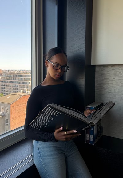 Joven mujer con gafas está junto a una ventana leyendo un libro grande y negro en una habitación moderna con edificios de la ciudad visibles afuera.