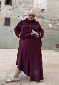 Άτομο που φοράει μπεζ μπέρε, μεγάλα γυαλιά, μπορντό φλις μπουφάν, φούστα σε τόνους που ταιριάζουν, λευκά αθλητικά παπούτσια και κρατάει μια μικρή καρό τσάντα.