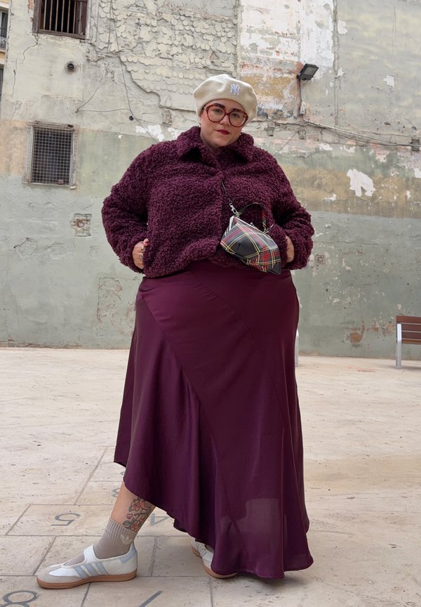 Osoba ubrana w beżowy beret, duże okulary, bordową polarową kurtkę, pasującą spódnicę, białe trampki i trzymająca małą kraciastą torebkę.