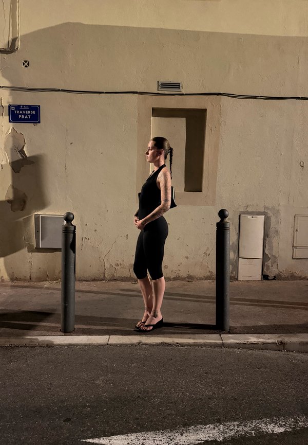 Giovane donna con capelli intrecciati e tatuaggi in piedi tra due paletti neri su un marciapiede urbano poco illuminato di notte.