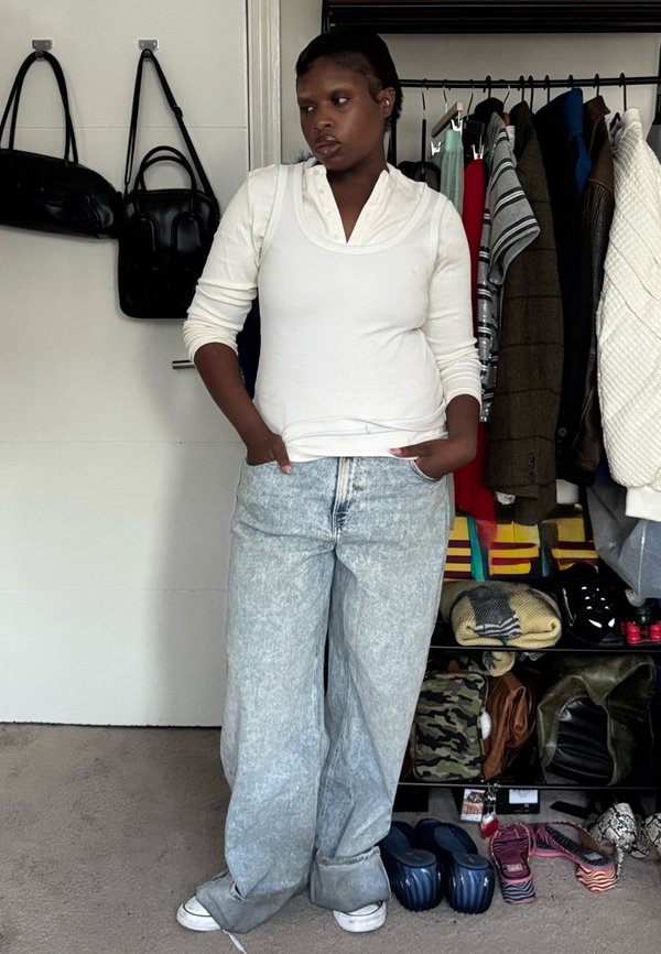 Person i hvid langærmet skjorte og brede acid-wash jeans står med hænderne i lommerne nær tøjstativ og skohylde.