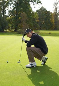 Moški v črni kapici in jakni se priklanja na zelenici za golf, drži golf palico in se pripravlja na udarec tik ob golf žogi, v ozadju so drevesa.