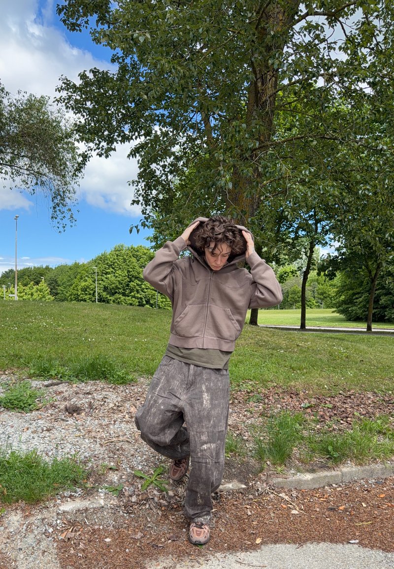 Personne aux cheveux bouclés portant un sweat à capuche marron et un pantalon ample se tient dehors sous un grand arbre sur un sol herbeux par une journée partiellement nuageuse.