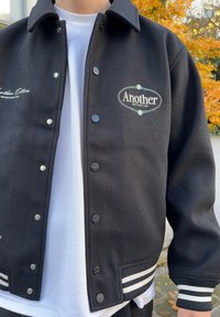 Mustast värvi ülikoolijakk valgete triibuliste mansettidega. Omab "Another Cotton Lab" kirjaga tikitud logot. Flanelltakistus, surunupud ja krae kujundus.