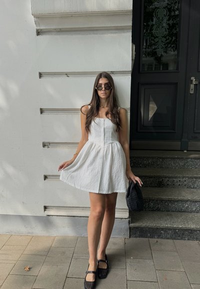 Vestido mini blanco texturizado con escote cuadrado y botones en el frente. Llevado con bailarinas negras tipo Mary Jane y gafas de sol, sujetando un bolso negro.
