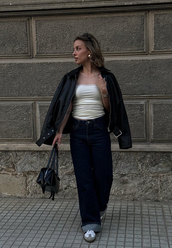 Femeie purtând o jachetă neagră de piele peste un top alb, blugi evazați închisi la culoare, ținând o geantă neagră, mergând pe o trotuar pavat lângă un zid de piatră.