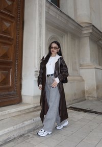 Long manteau en cuir brun sur un haut blanc, jean gris à jambes larges et baskets noires et blanches. Les accessoires incluent des lunettes de soleil et des colliers superposés.