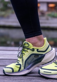 Neon sárga futócipő hálós anyagból, fényvisszaverő részletekkel és mintás talppal. A fűzők fekete alapon zöld mintázattal díszítettek.