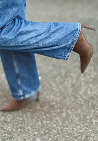 Chaussure à talon haut en daim marron associée à un jean bleu, avec une coupe décontractée et un ourlet effiloché, présentée en angle sur une surface texturée.