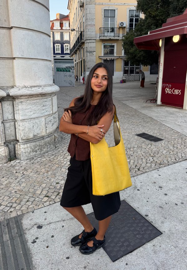 Mujer con cabello largo y oscuro está de pie en una calle adoquinada cerca de edificios, lleva un top marrón, pantalones cortos negros, zapatos negros y lleva un gran bolso amarillo.