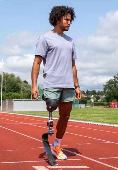 Pierna prostética con una estructura de fibra de carbono negra y forma curva, utilizada por un hombre con una camiseta gris claro y pantalones cortos verdes en una pista de atletismo.
