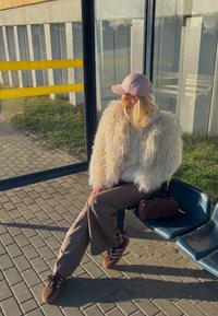 O persoană stă pe un scaun albastru din autobuz, purtând o jachetă albă pufoasă, un hanorac bej, pantaloni maro și adidași maro cu dungi roz. Lângă ea se află o geantă închisă la culoare.