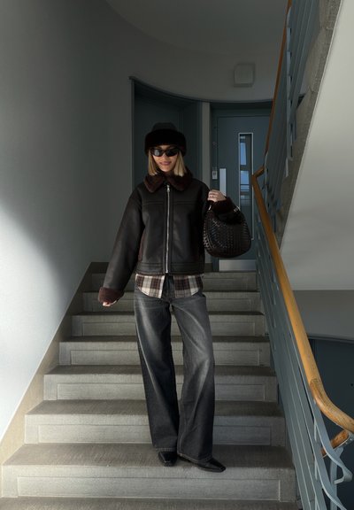 Chaqueta de cuero sintético negra con cuello marrón de pelo, camisa a cuadros, vaqueros anchos oscuros y bolso negro tejido, de pie en una escalera.