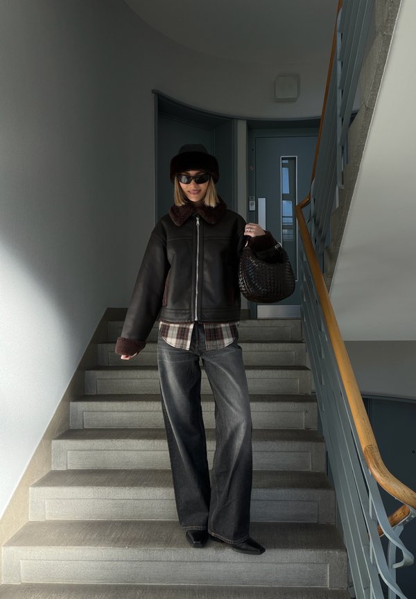 Svart jacka i konstläder med brun lurvig krage, rutig skjorta, vida mörka jeans och en svart flätad handväska, stående på en trappa.