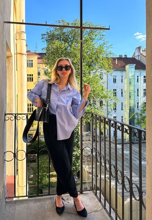 Camisa azul claro de botones, pantalones negros, bolso negro y tacones puntiagudos, de pie en un balcón con vegetación y edificios de fondo.