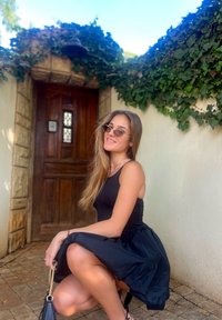 Μαύρο φόρεμα με εφαρμοστό τοπ και φουσκωτή φούστα. Μακριά μαλλιά, καθισμένη απέναντι από μια πέτρινη τοιχοποιία με κισσό. Τα αξεσουάρ περιλαμβάνουν γυαλιά ηλίου και μια μικρή μαύρη τσάντα.