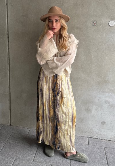 Blusa de un beige claro y texturizado con mangas voluminosas, combinada con una falda maxi fluida de tinte anudado en tonos amarillos y morados. Zapatos de lona oliva.