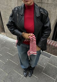 Czarna skórzana kurtka, czerwony golf, beżowe rękawiczki, szerokie dżinsy z niebieskiego denimu oraz czerwono-biały pasiasty szalik trzymany w ręce.