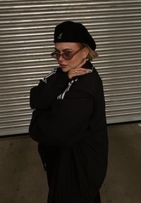 Μαύρο φαρδύ μπουφάν με λευκές ρίγες, μαύρο μπερέ με λογότυπο, γυαλιά ηλίου με απόχρωση και έντονα δαχτυλίδια. Στέκεται μπροστά από μια μεταλλική επιφάνεια.