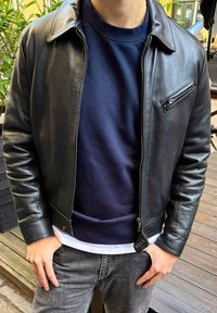 Czarna skórzana kurtka z zamkiem błyskawicznym z przodu, lewą kieszenią na piersi i ściągaczami przy mankietach, noszona na granatowej bluzie i szarych dżinsach.
