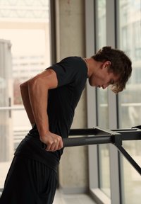 Atletisk person utfører dips på en svart metall dip-stasjon med teksturerte grep og en industriell bakgrunn med store vinduer.