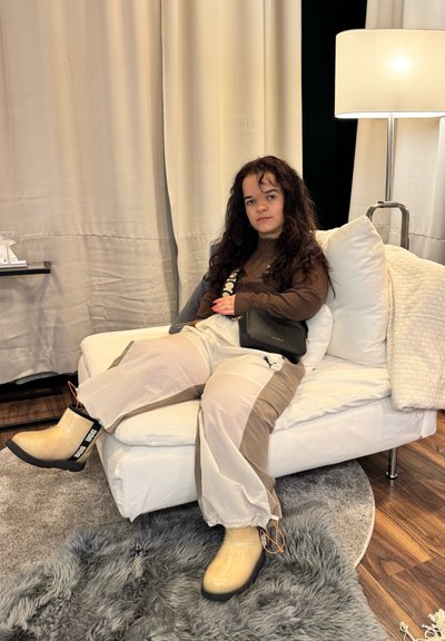 Botas de goma beige con una correa negra, pantalones de un ligero tono gris y marrón con un estampado, y una camiseta marrón. Sentada en un sofá blanco con almohadas.
