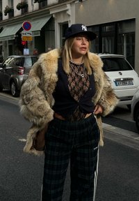 Žena v béžovom kabáte s kožušinou, pod ktorým má čierny čipkovaný top, tmavých károvaných nohaviciach s bielymi pruhmi a čiernej šiltovke s logom.