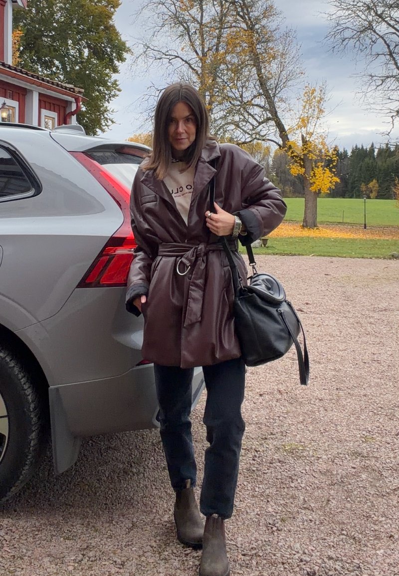 Bruine leren trenchcoat met een riem, lichte trui, zwarte tas, donkere jeans en bruine enkellaarsjes, staand naast een grijze SUV in een buitenomgeving.