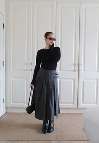 Top preto de mangas compridas, saia midi cinza xadrez com pregas e fivelas, botas pretas de tornozelo e pequena bolsa preta contra um fundo claro.