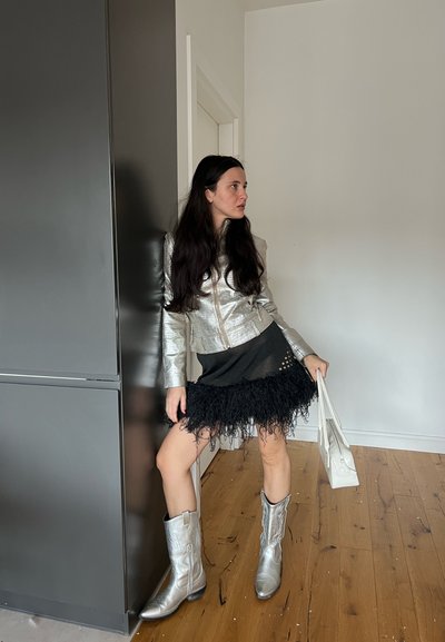 Chaqueta metálica plateada, falda negra con flecos, botas de cowboy plateadas y un bolso blanco. Texturas suaves con materiales y patrones mezclados.