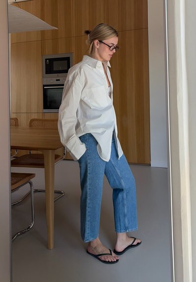Mujer con camisa blanca, vaqueros azules, chanclas negras y gafas, de pie en una cocina moderna con armarios de madera y mesa de comedor.