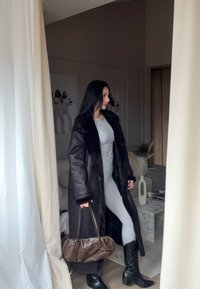 Palton lung, maro închis, cu guler de blană, costumul gri, cizme înalte negre și o geantă mică maro. Fundalul prezintă o cameră de culoare deschisă.