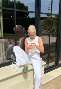 Λευκή ριγέ μπλούζα χωρίς μανίκια με το κείμενο "LUCKY", λευκά παντελόνια με φαρδιά γραμμή και μωβ ρίγες, πράσινες σαμπάνιες, λευκή τσάντα και διαφανές ποτό.