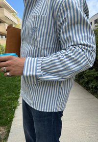 Pessoa vestindo uma camisa listrada azul e branca e jeans escuros, segurando uma pasta de couro castanha e um copo com tampa azul ao ar livre.