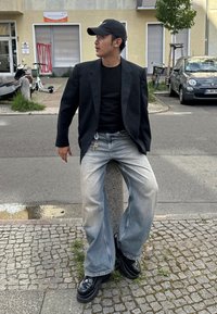 Črni blazer s pikastim vzorcem preko črnega majice, v kombinaciji z ohlapnimi svetlo modrimi jeansom in debelimi črnimi čevlji. Ključna veriga vidna na pasu.