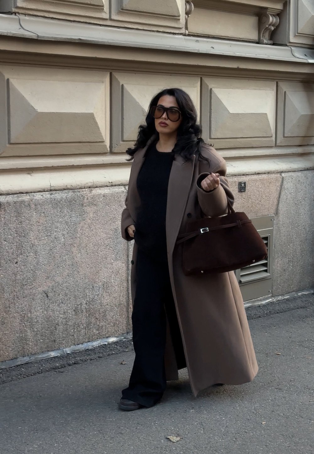 Palton lung maro peste o ținută neagră, cu o geantă maro structurată. Paltonul are o textură netedă și nasturi mari, completând pantofii negri.