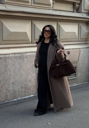 Abrigo largo marrón sobre un conjunto negro, con un bolso marrón estructurado. El abrigo tiene una textura suave y grandes botones, que complementan los zapatos negros.