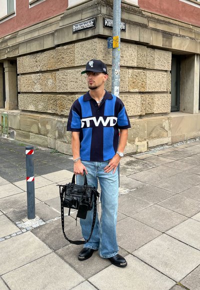 Camisa polo de manga corta con rayas azules y negras, gran logotipo "GTWD", jeans de color azul claro, llevando un bolso negro texturizado, de pie sobre una acera.