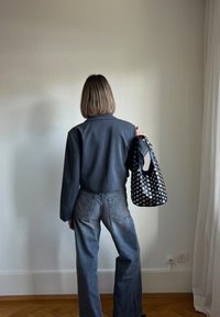 Osoba oblečená v tmavě šedém saku a volných šedých džínách, držící černou tašku s kovovými hroty na texturovaném pozadí.