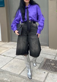 Púrpura blús satinada con mangas largas, corbata negra, culottes de mezclilla de cintura alta y botas plateadas con estampado de serpiente hasta la rodilla. Clutch negro en la mano.