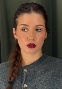 Grijze gebreide kleding met knopen, gecombineerd met een ingevlochten haarstijl. Heeft dieprode lippenstift en een septumpiercing. Groene achtergrond.