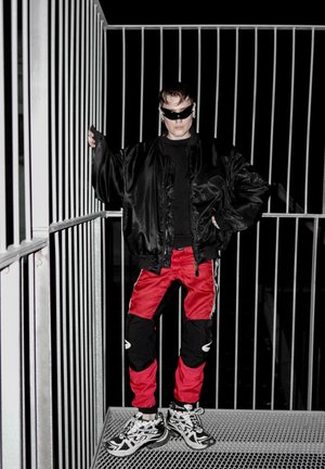 Chaqueta bomber de satén negra, camiseta negra, pantalones deportivos rojos y negros con rayas blancas, gafas de sol negras de gran tamaño y zapatillas deportivas con un diseño grueso.