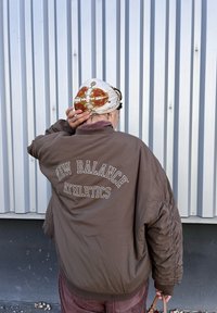 Καφέ τζάκετ βομβαρδιστικού με ribbed μανίκια και γιακά, διαθέτοντας το κείμενο "NEW BALANCE ATHLETICS" στην πλάτη. Φοριέται με ένα χα patterned κασκόλ.