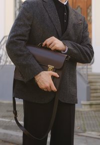 Mallette en cuir marron avec poignée supérieure et bandoulière réglable, dotée d'un fermoir en métal et de détails de couture, tenue par une personne portant une veste grise.