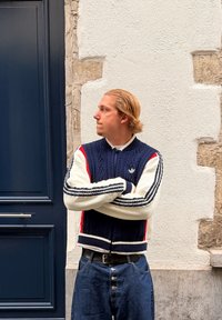 Tmavomodrý a krémový pletený sveter so červenými akcentmi, ktorý má textúrovaný vzor, žebrované manžety a golier, nosený cez biele tričko a džínsy.