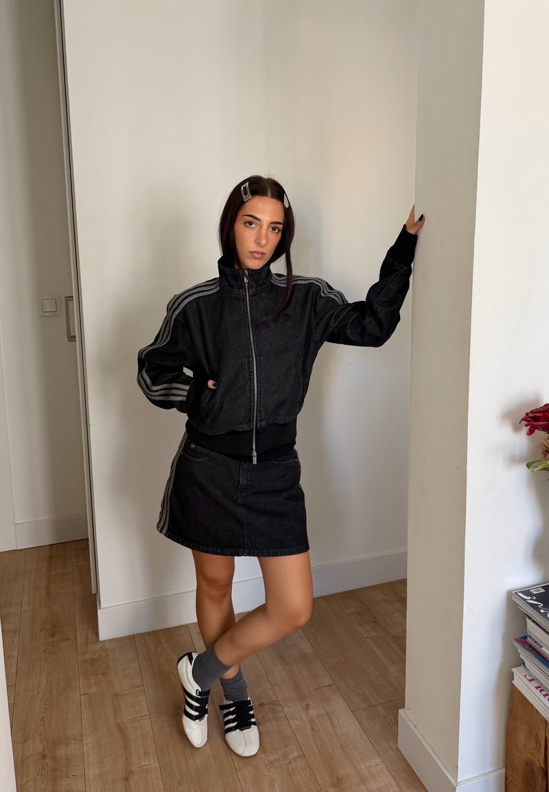Jeune femme appuyée contre un mur, portant une veste noire zippée, une jupe noire, des chaussettes grises et des baskets blanches avec des rayures noires.