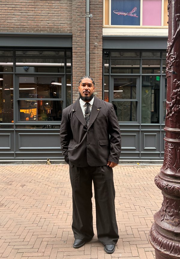 Man with braided hair and beard wearing a dark oversized suit, standing on a brick street in front of a building with large windows.
