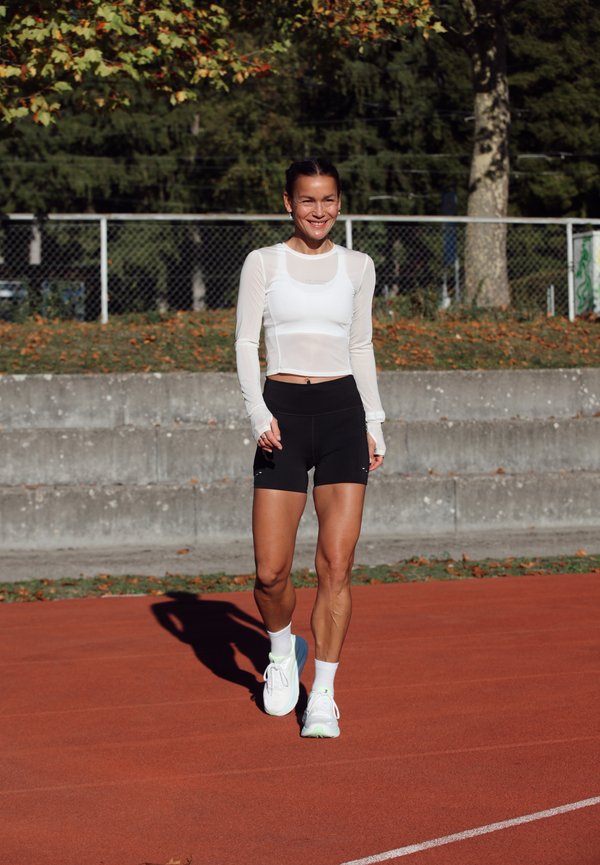 Une femme souriante en tenue de sport traverse une piste de course rouge, avec des arbres et un grillage derrière elle.