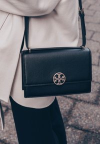 Crna kožna torba preko tijela s glatkom teksturom, koja ima okrugli zlatni logo kopču i preklopno zatvaranje.