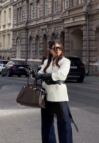 Weißes Sakko, schwarze Lederhandschuhe, dunkelblaue Hose mit Spitzenbesatz, braune Handtasche mit goldener Hardware und große dunkle Sonnenbrille. Urbaner Hintergrund.